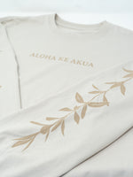 A close-up of a dust long-sleeve Tshirt with 'ALOHA KE AKUA' printed in tan across the chest and a matching tan maile print running down both sleeves.